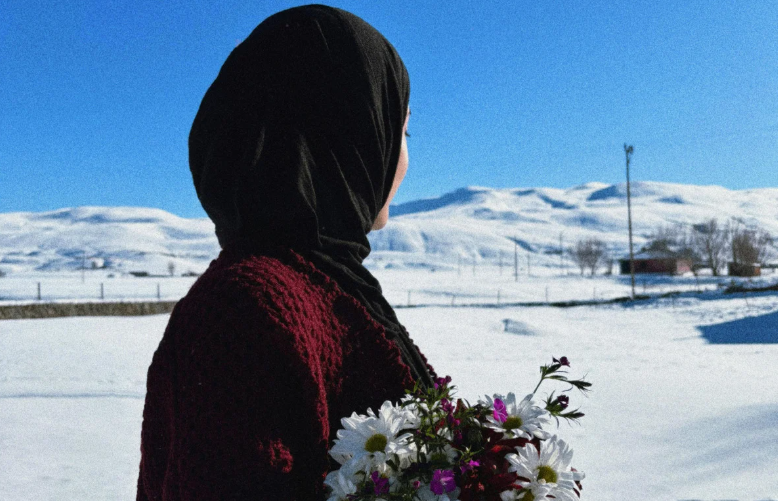 Femme en hijab de profil tenant un bouquet de fleurs dans les mains sur un fond de montagne enneigée.